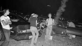 A group of young people stand and talk near parked cars at night. One person is facing the camera and smiling, while others are talking or leaning on cars. Smoke rises from one car, and the scene has a casual, candid atmosphere.