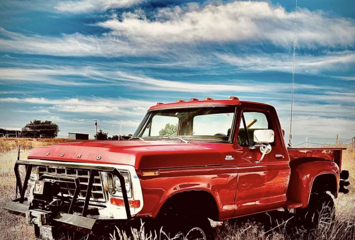 A vintage red Ford pickup truck is parked in a grassy field under a blue sky with wispy clouds. The truck appears weathered but well-maintained, with some grass and plants in the foreground.