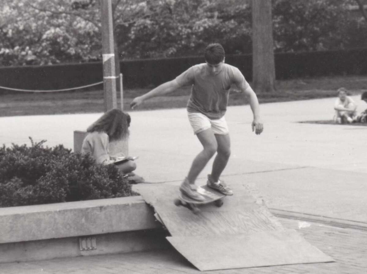 A young man skateboards down a wooden ramp in an outdoor area, while a woman sits nearby on a ledge, writing or drawing. Trees and people sitting on the ground are visible in the background. The photo is in black and white.