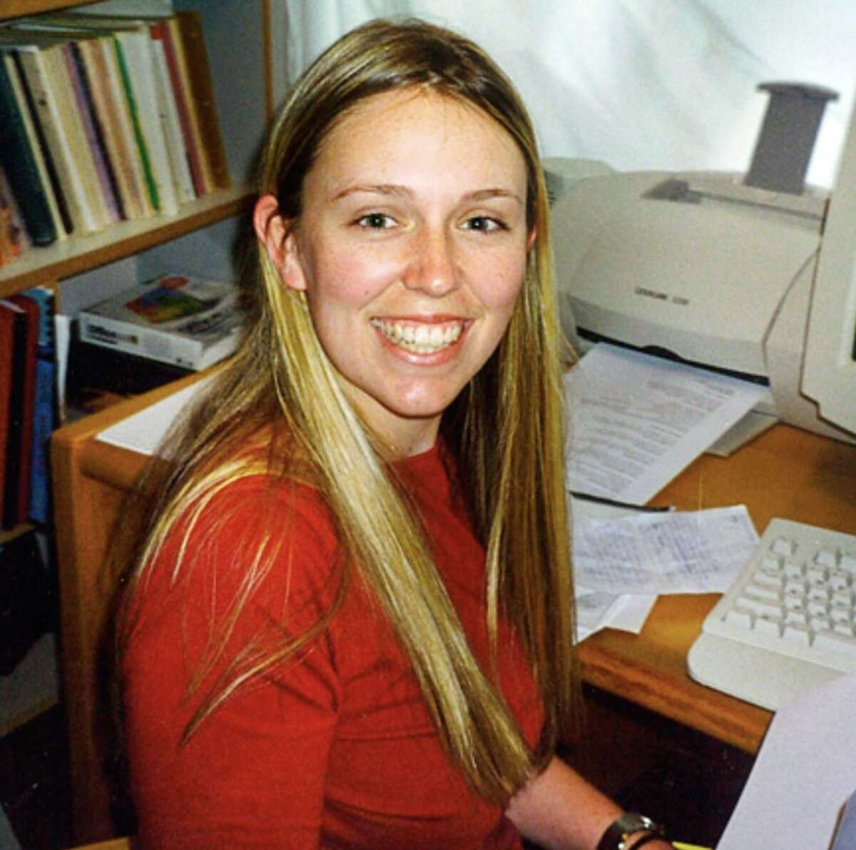 A smiling woman with long blonde hair, wearing a red top, sits at a desk with papers, a computer keyboard, and bookshelves in the background.