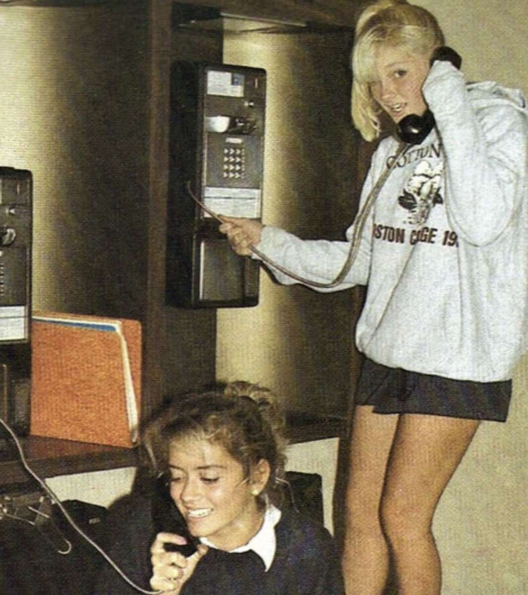 Two young women use payphones. One is standing and wearing a light sweatshirt, holding the phone and a wire, while the other is crouched below, smiling and talking on a phone. A bright binder is on the shelf beside them.