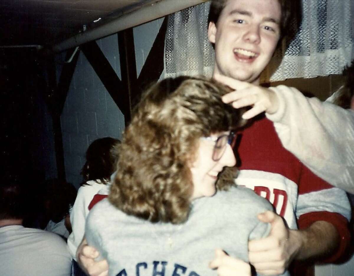 A young man smiles while playfully patting a young woman's head from behind. Both appear happy, with the woman wearing glasses and a grey sweatshirt, and the man in a red and white shirt. Indoors with others in the background.