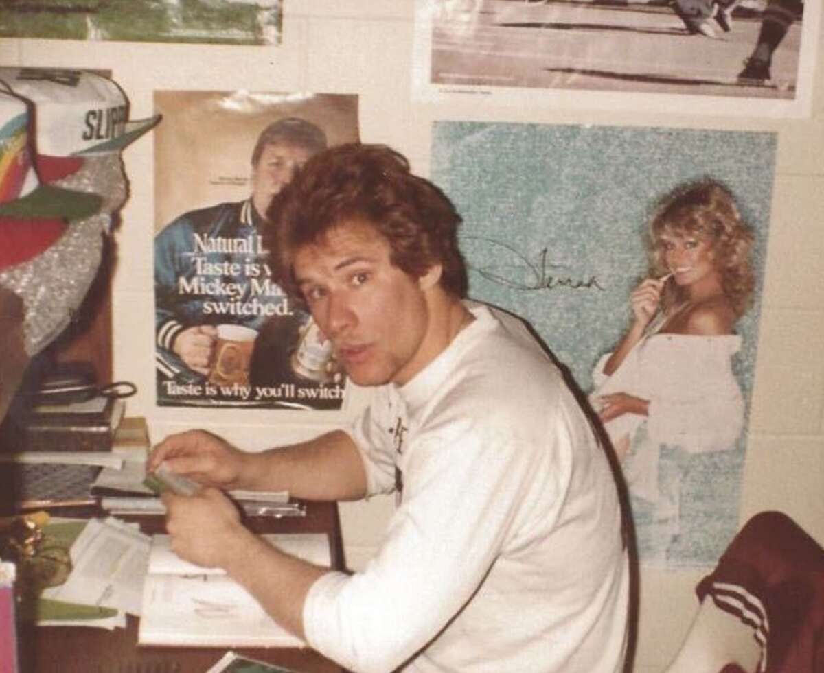 A man with brown hair in a white shirt sits at a cluttered desk, turning to look at the camera. Behind him are posters, including one of a woman in white and another of a man holding a mug.