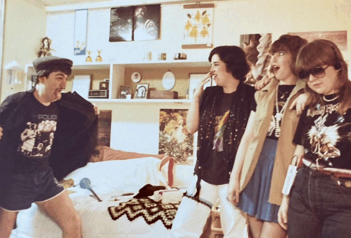 Four young people in a retro bedroom, three standing and laughing near the bed while one person playfully flashes their Police t-shirt, sticking out their tongue. The room is decorated with posters and various items on shelves.