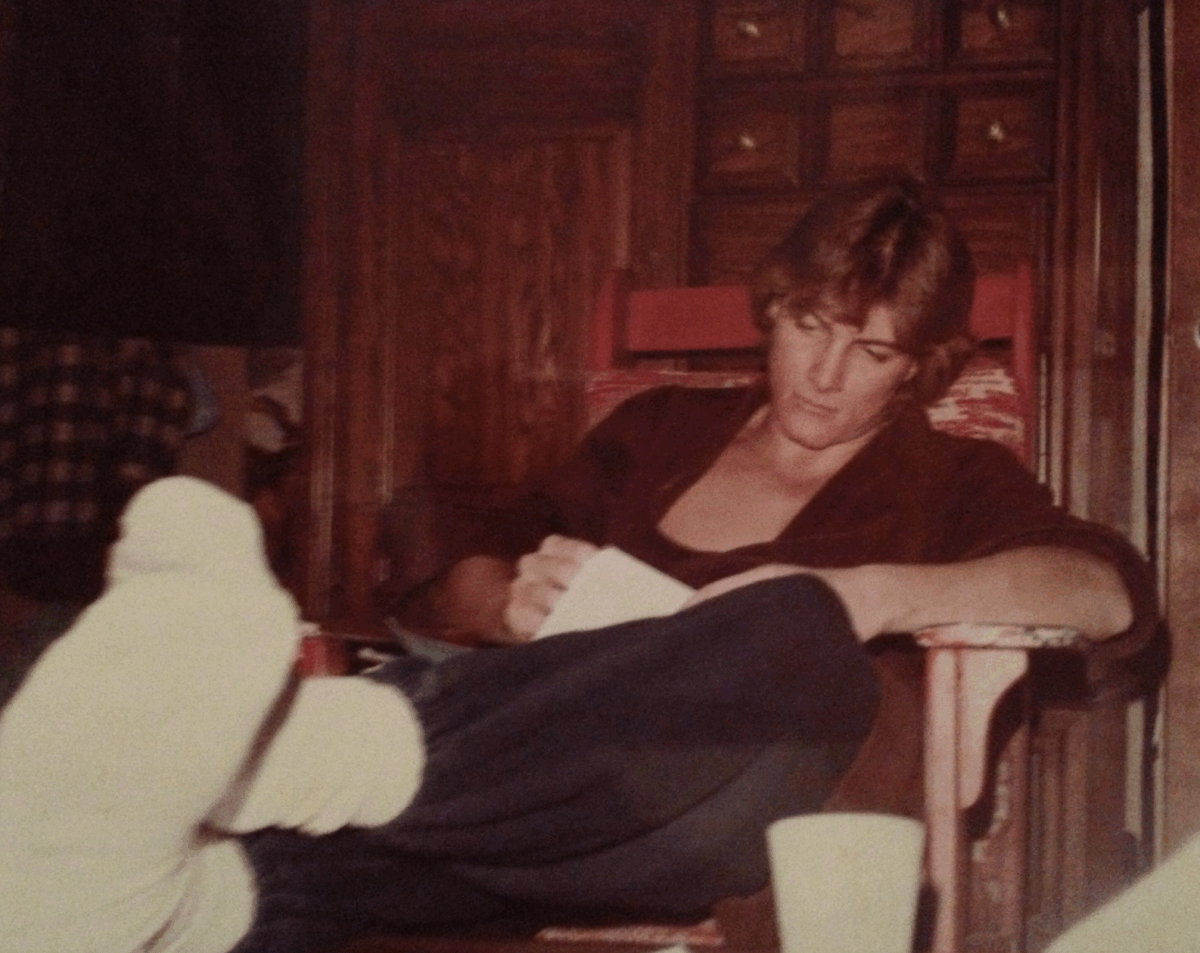 A young person with light brown hair sits in a chair indoors, dressed in dark pants, a dark top, and white socks, holding a piece of paper and appearing to write or read, with a wooden cabinet in the background.