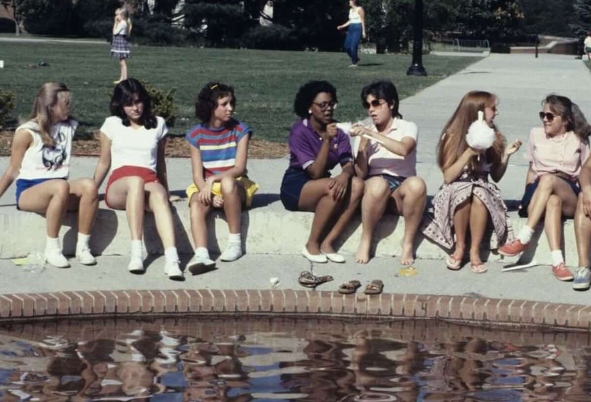 Seven young women sit on the edge of a fountain, chatting, eating, and relaxing on a sunny day. Shoes are off, and their reflections are visible in the water. A grassy park is in the background with another person walking.