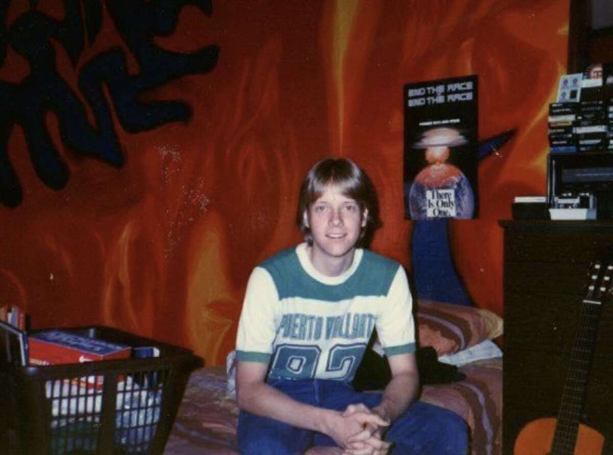 A young person with light brown hair sits on a bed in a room with bright red and orange walls. Behind them is a poster, books, a guitar, and a laundry basket. The person is smiling and wearing a "Puerto Vallarta 82" shirt.