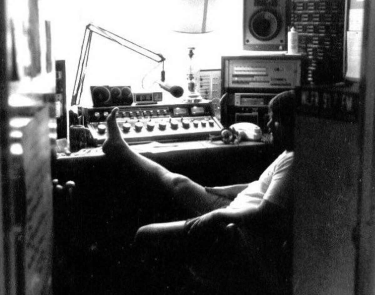 A person sits in a dimly lit radio studio, leaning back with feet up on the console, surrounded by audio equipment, dials, and a microphone, with light coming from a lamp in the background.