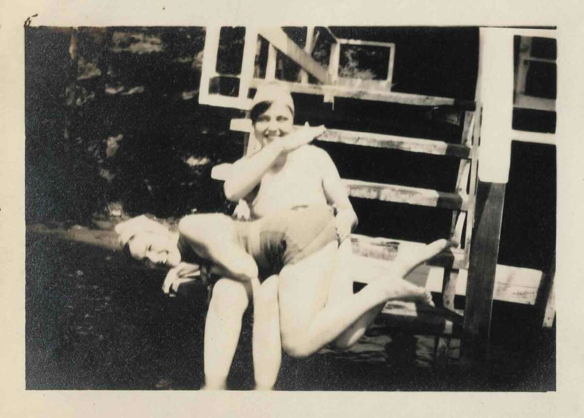 Two people are playfully posing outside in front of wooden steps. One person is sitting and pretending to spank the other, who is bent over their lap, both laughing. The photo is black and white and slightly blurry.