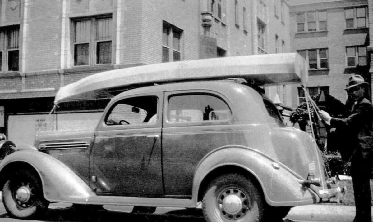 A black-and-white photo shows an old-fashioned car parked on a city street, with a long canoe strapped to its roof. A man in a suit and hat stands beside the vehicle, holding the canoe.