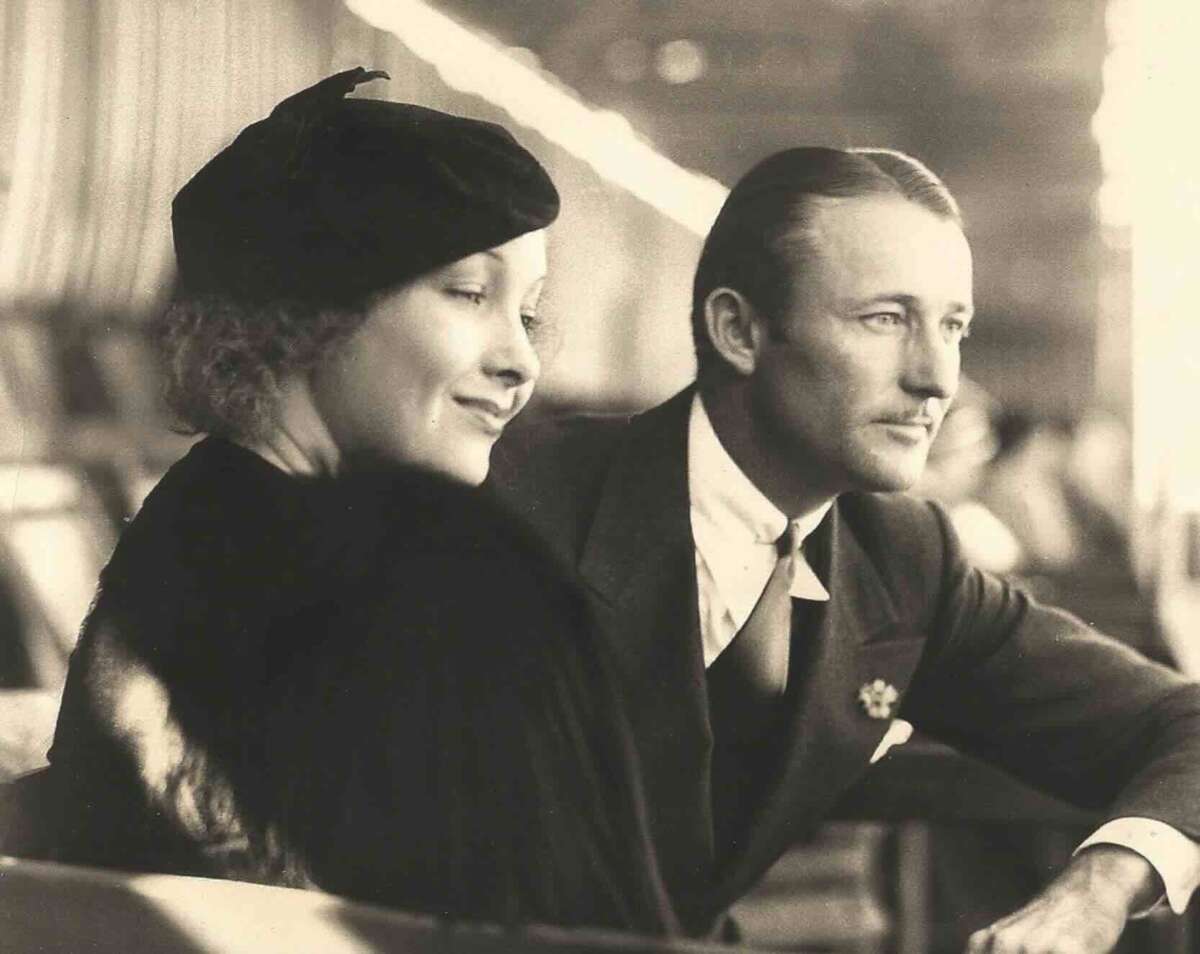 A black-and-white photo of a woman and a man in formal attire sitting side by side. The woman wears a hat and fur, smiling softly, while the man, in a suit, gazes thoughtfully into the distance.
