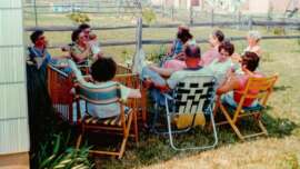 A group of people sit in lawn chairs arranged in a circle outdoors on grass, chatting and holding drinks. A baby is present. There is a wooden fence and a swing set in the background. The scene is relaxed and sunny.