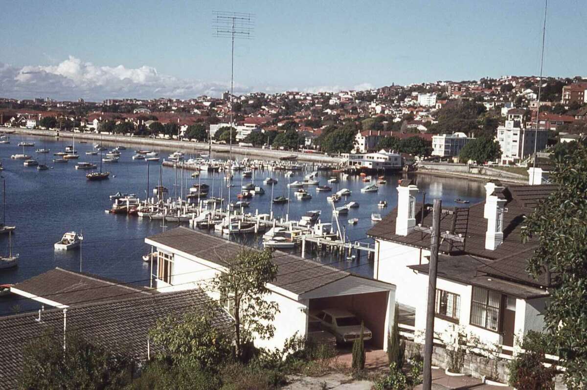 A coastal suburb with many boats moored in a bay, residential houses in the foreground, and a hillside of homes and trees in the background under a clear blue sky.