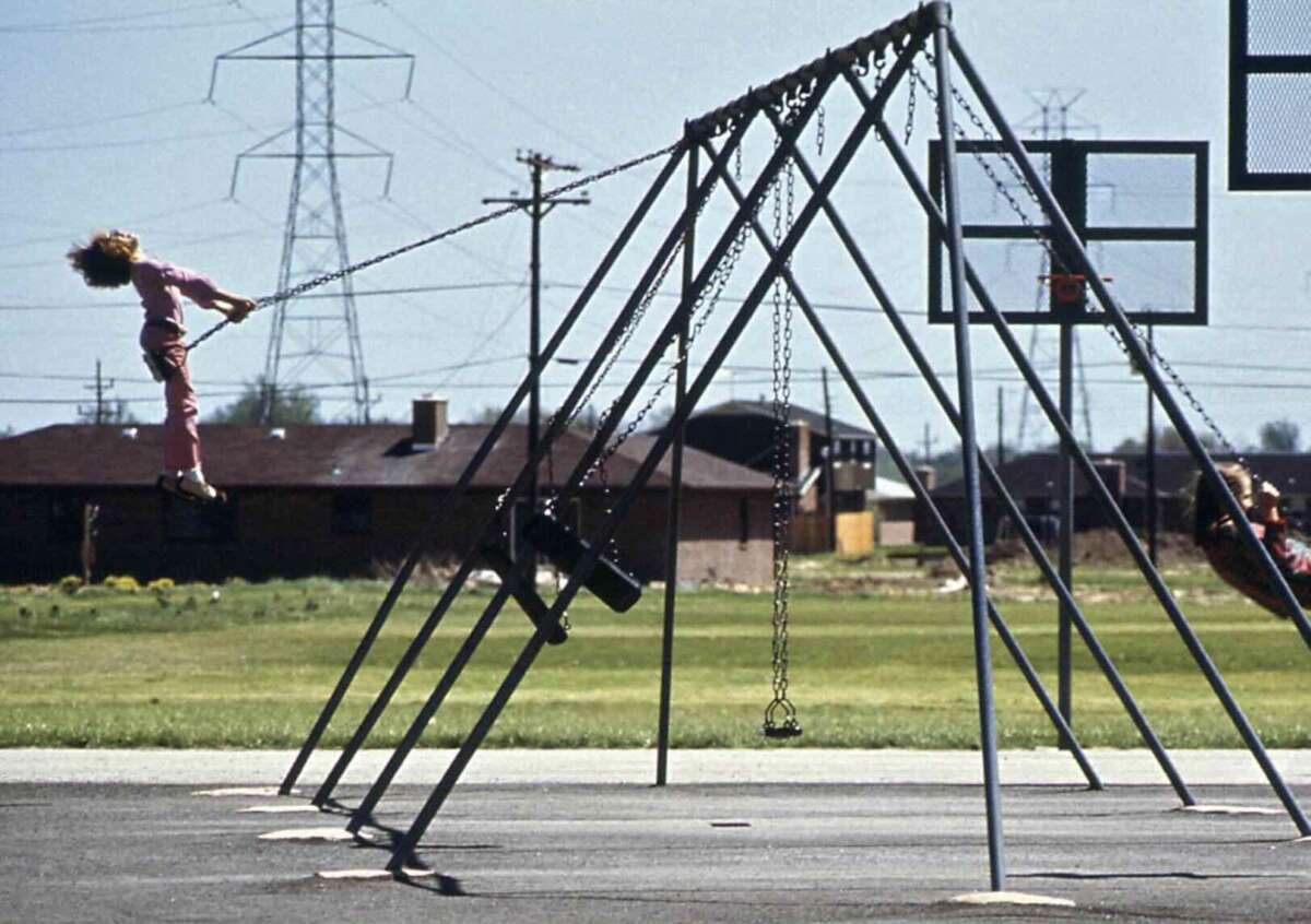 A child in pink pants swings high on a playground swing set, with a grassy field, basketball hoop, power lines, and buildings in the background.