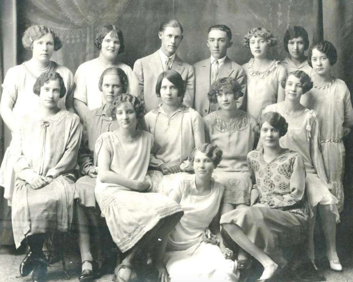 A black-and-white vintage group portrait of thirteen women and two men in 1920s-style clothing, seated and standing in rows, posing formally for the camera against a plain studio backdrop.