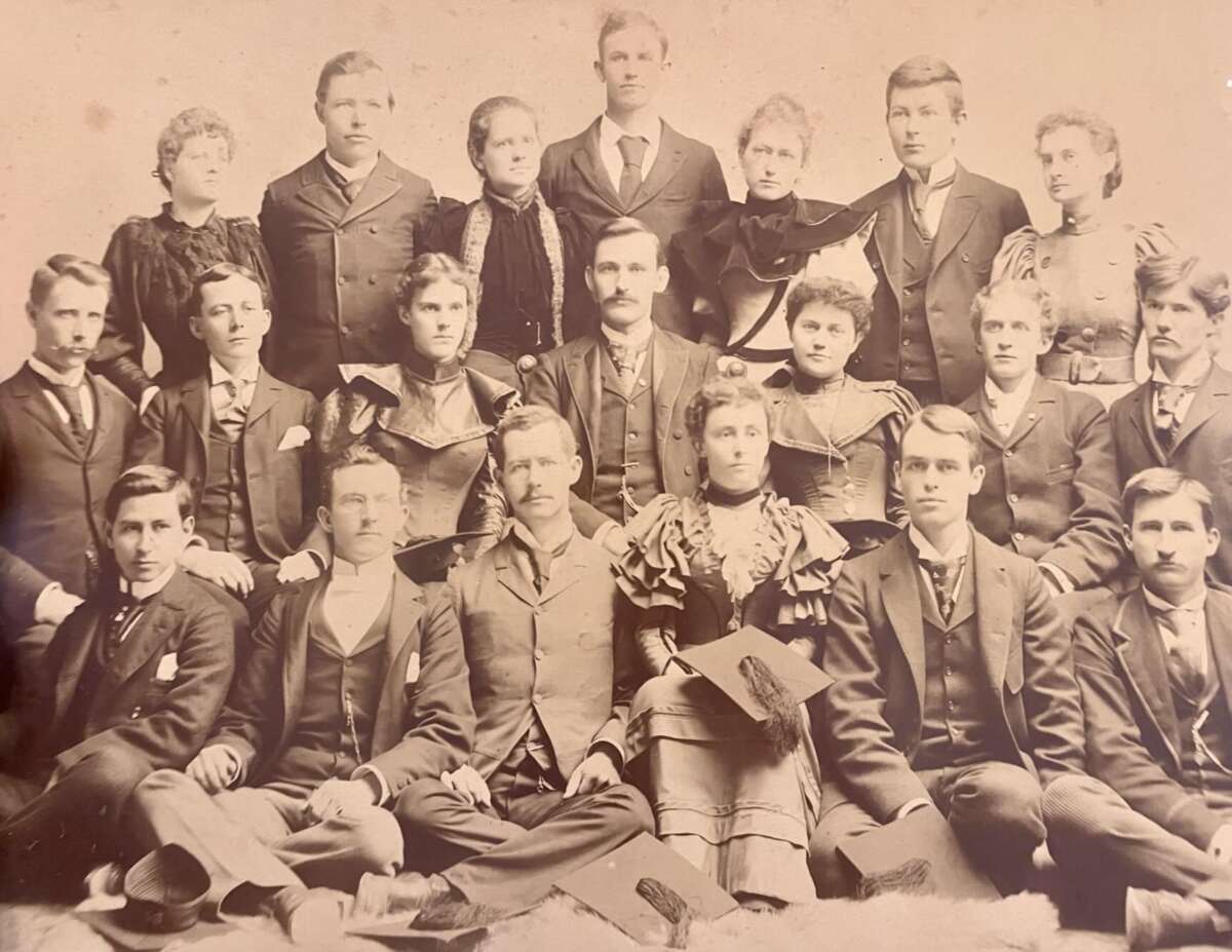 A sepia-toned vintage photograph shows a group of nineteen men and women in formal Victorian-era clothing, posed in three rows and looking at the camera with serious expressions.