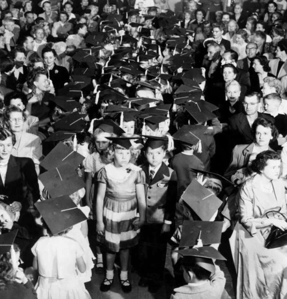 A black-and-white photo shows a large group of young children in graduation caps, standing among seated adults at a crowded ceremony. Two children in front face the camera while others look in various directions.