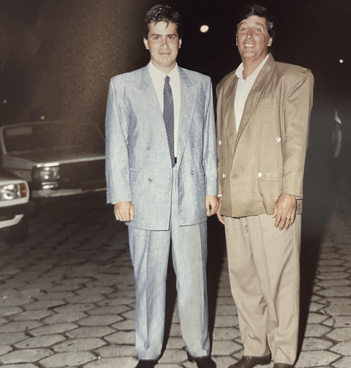 Two men in suits stand side by side and smile at night on a cobblestone street, with a parked car and streetlights visible in the background.