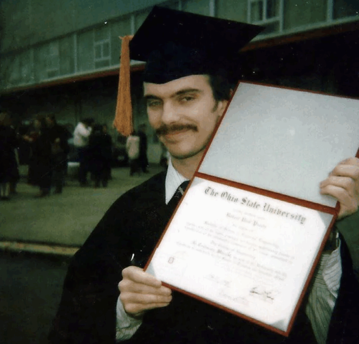 A person in a graduation cap and gown smiles while holding up an open Ohio State University diploma folder outdoors, with people and a building visible in the background.