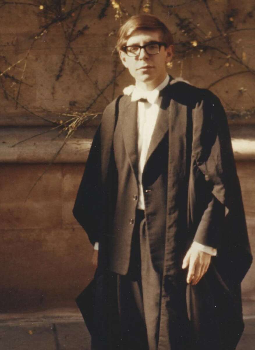 A young man in glasses wears academic robes and stands outdoors in front of a stone wall with some vines. Sunlight casts shadows on the wall behind him.
