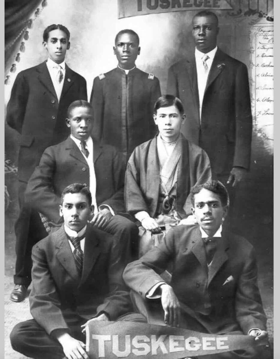 Seven young men in formal suits pose for a black-and-white portrait. They are seated and standing in two rows, holding a banner that reads "TUSKEGEE." The background includes draped fabric and partial signage.
