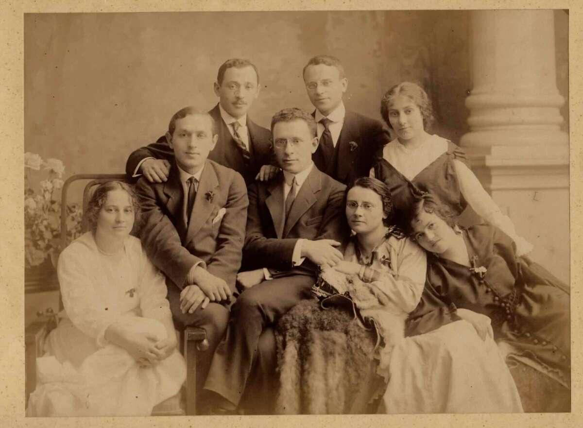 Vintage sepia photo of five men in suits and three women in dresses, seated and standing closely together, posing formally in front of a classical column backdrop.