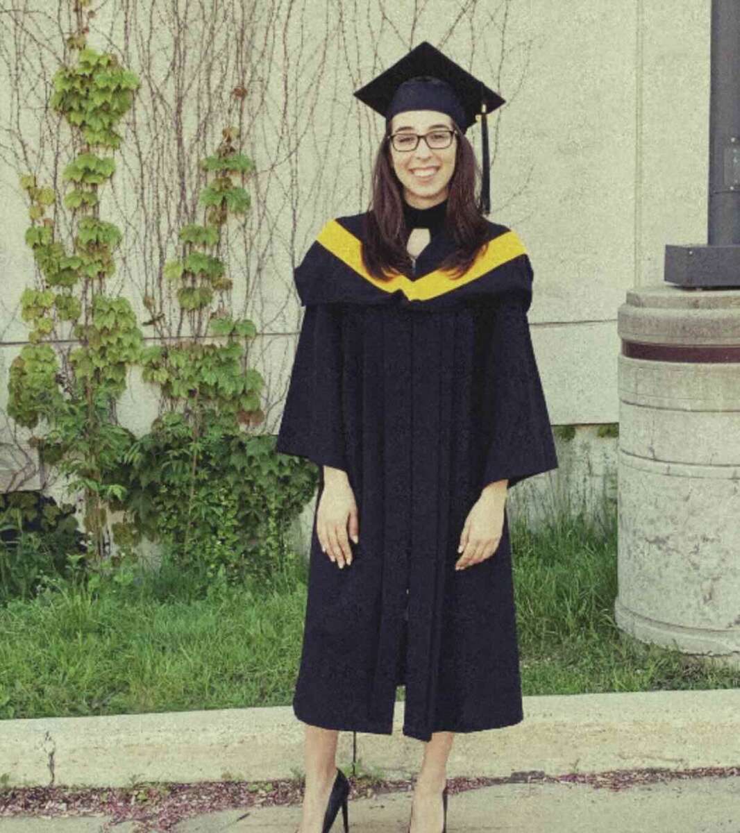 A person wearing a black graduation gown, cap, and glasses stands smiling outdoors in front of a concrete wall with some greenery.