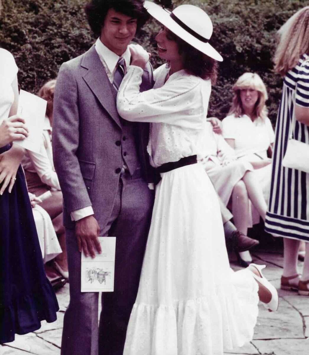 A young man in a gray suit stands smiling next to a woman in a long white dress and wide-brim hat, who is adjusting his tie and playfully lifting her leg. Several people in light-colored outfits sit in the background outdoors.