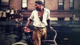 A young man with a white shirt and pendant stands beside a bicycle on a wet city street, with children and adults gathered in the background near a brick building.