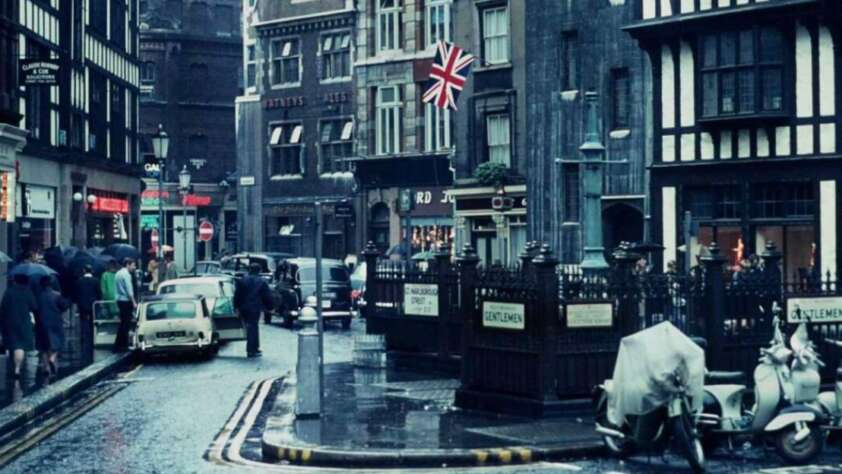 A busy, rainy street scene in London with people walking, vintage cars parked, scooters, a British flag, and historic black-and-white buildings. Signs indicate entrances to public toilets.
