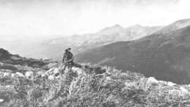 A person with a rifle stands among rocks and brush on a mountain slope, overlooking a vast landscape of forested hills and distant peaks under a hazy sky.