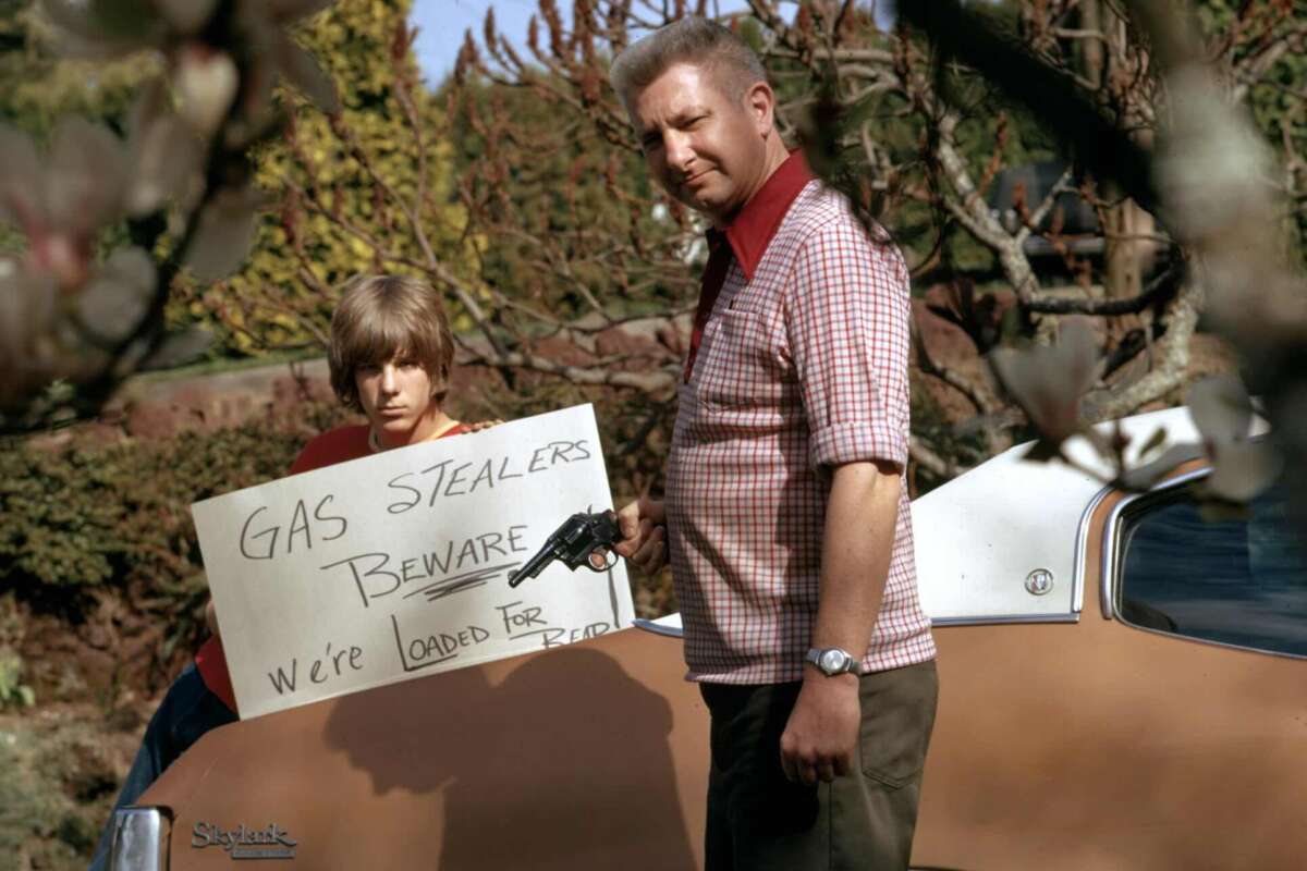 A man holds a revolver and a handwritten sign that reads, "Gas stealers beware, we’re loaded for bear," while a boy stands beside him near a car. Trees and bushes are in the background.