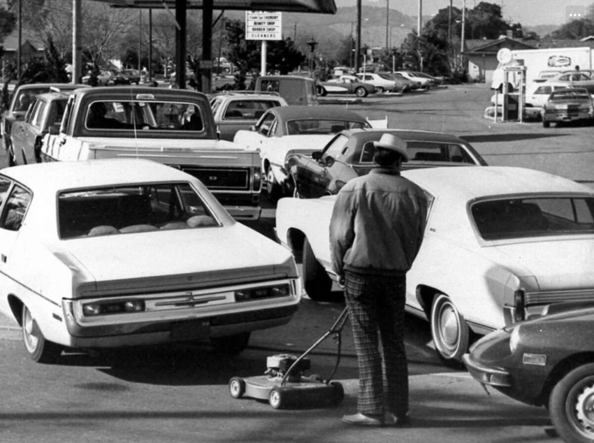 A man in plaid pants stands with a lawn mower next to a line of cars at a gas station, suggesting a fuel shortage. The scene appears to be from the 1970s based on the cars and fashion.
