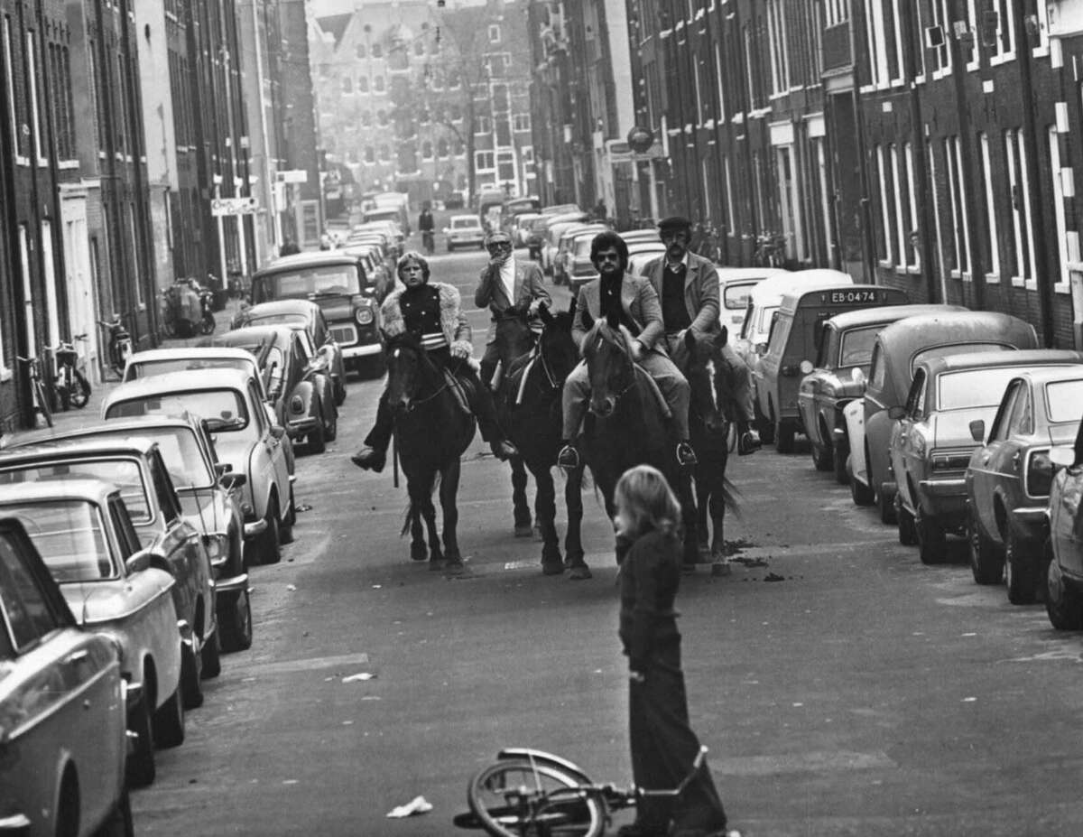 Three people riding horses and a fourth person on foot move down a narrow city street lined with parked cars. A child stands in the foreground near a fallen bicycle, looking at the group. The image is in black and white.