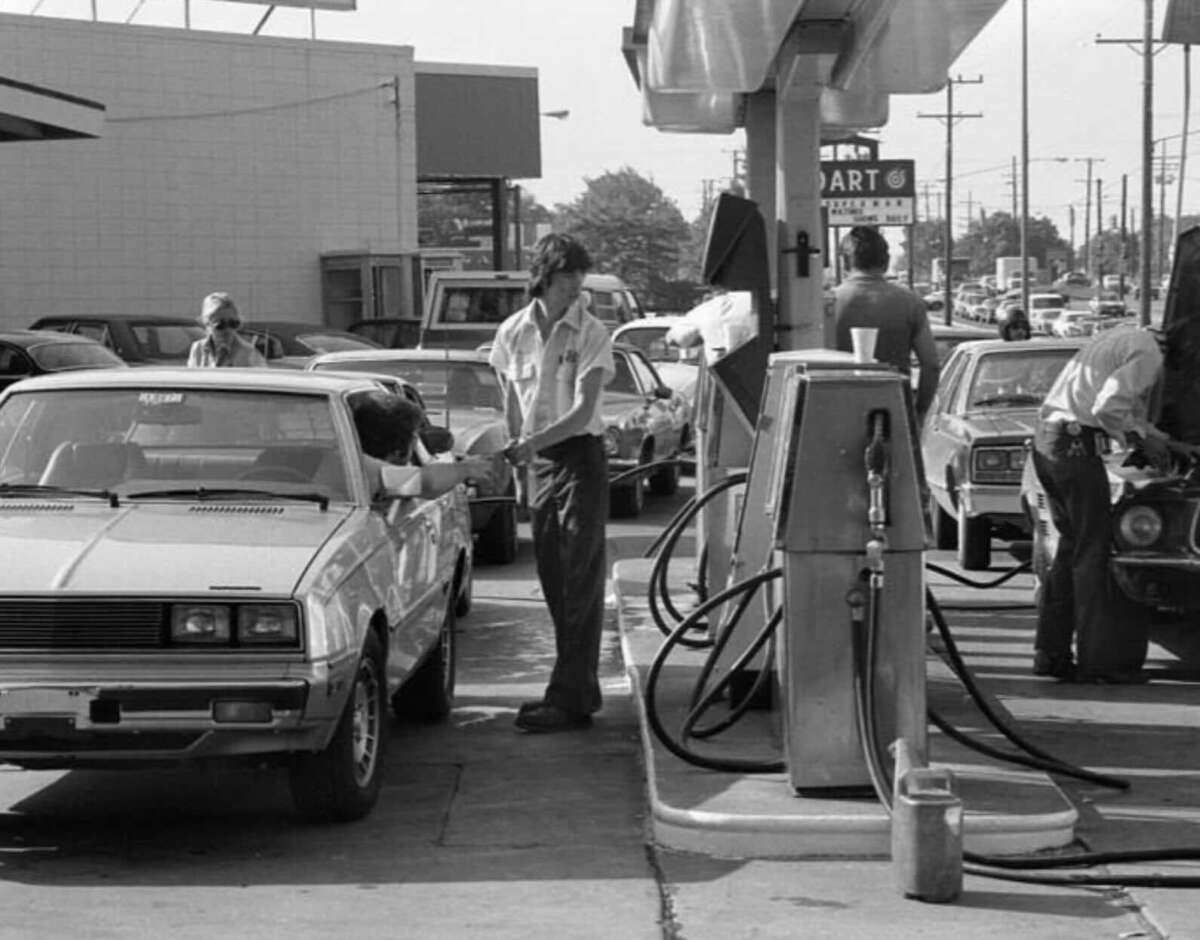 Black-and-white photo of people waiting in line at a busy gas station, fueling their cars. Multiple vehicles are parked by pumps, and attendants assist drivers on a crowded street.