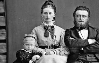 A vintage black-and-white portrait of a family: a woman sits in the center with a young girl in a dark dress on her left and a bearded man with folded arms on her right. The background shows wooden planks and a dark cloth.