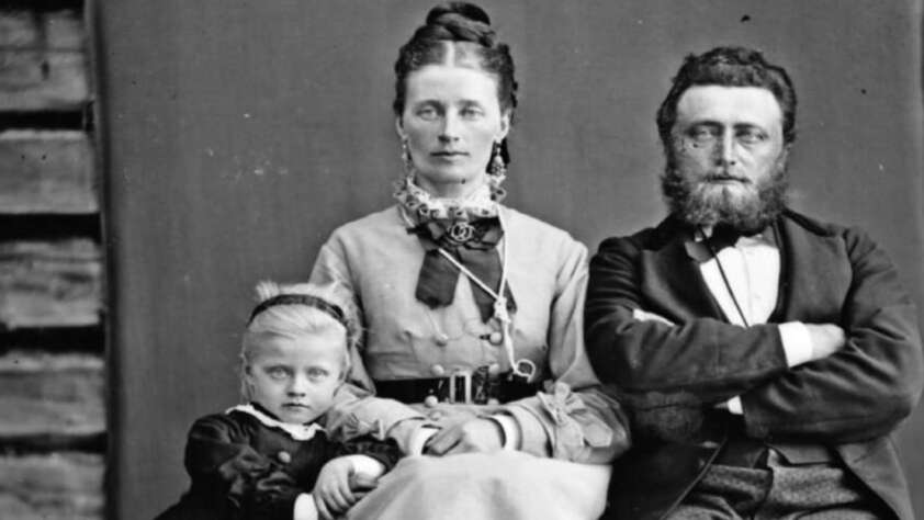 A vintage black-and-white portrait of a family: a woman sits in the center with a young girl in a dark dress on her left and a bearded man with folded arms on her right. The background shows wooden planks and a dark cloth.