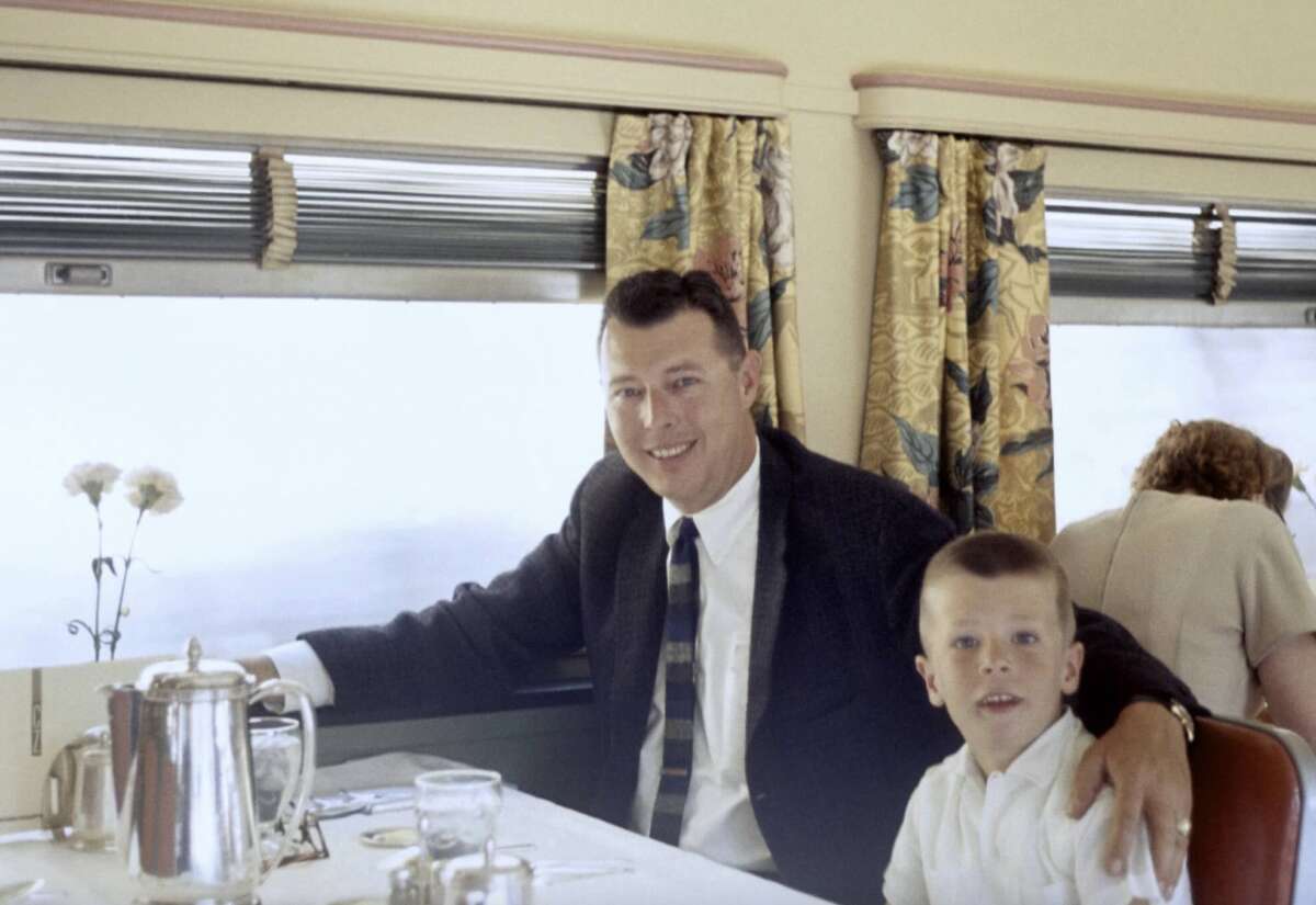 A man in a suit and a young boy sit together at a dining table on a train, smiling at the camera. Floral curtains and a window are in the background, with another person seated and turned away.