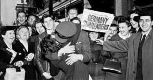 A joyful crowd celebrates in the street as a soldier kisses a woman; behind them, a person holds a newspaper with the bold headline "GERMANY SURRENDERS." People are smiling and laughing, clearly elated.