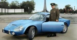 A man in a military uniform stands next to an open blue vintage sports car, parked outdoors near a monument with trees and several people in the background.