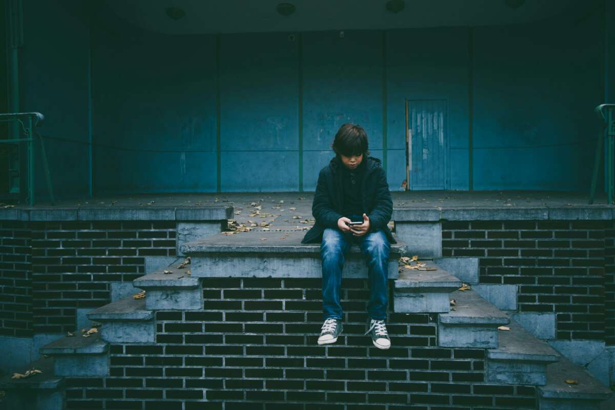 A boy in a dark coat and jeans sits alone on brick steps, looking down at a phone in his hands. Fallen leaves are scattered around him, and the background shows a blank blue wall and a closed door.