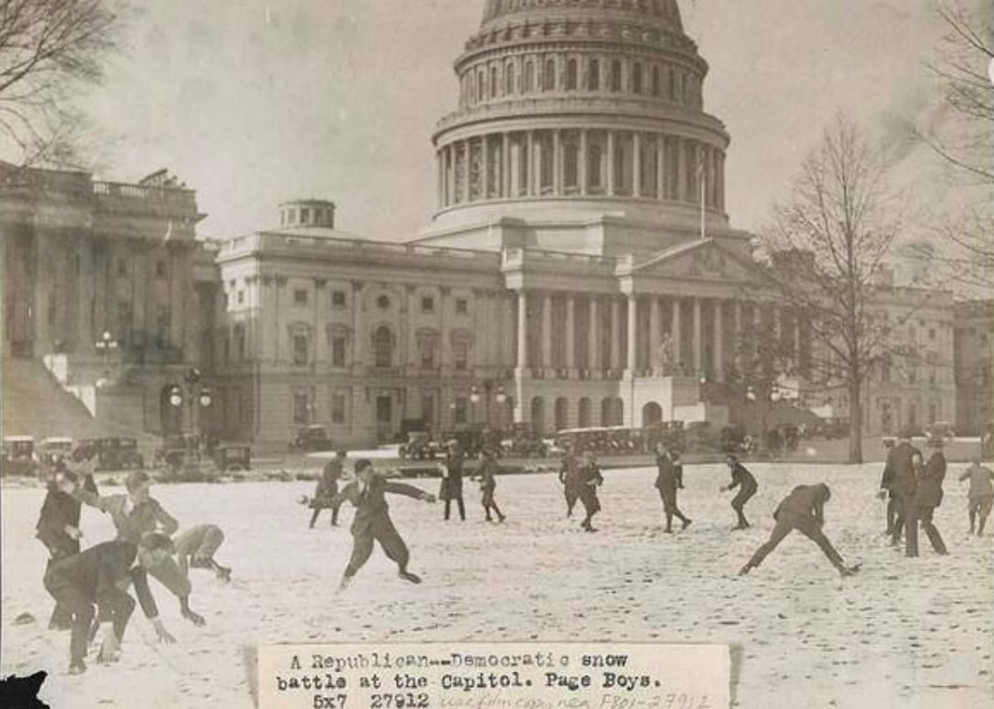 Children and adults have a snowball fight on the snowy lawn in front of the U.S. Capitol building in Washington, D.C., with vintage cars and trees in the background.