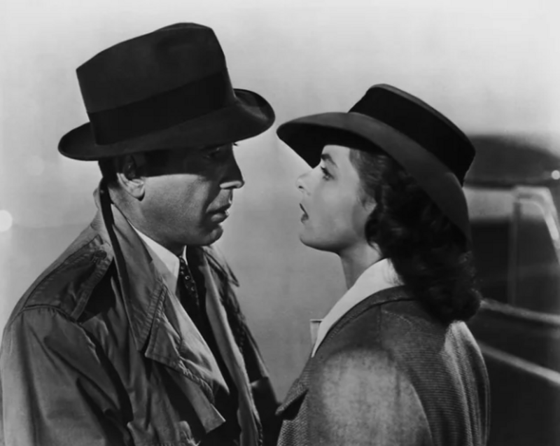 A man in a trench coat and fedora faces a woman wearing a wide-brimmed hat and coat. They stand close together, gazing at each other, in a misty, dramatic black-and-white setting.
