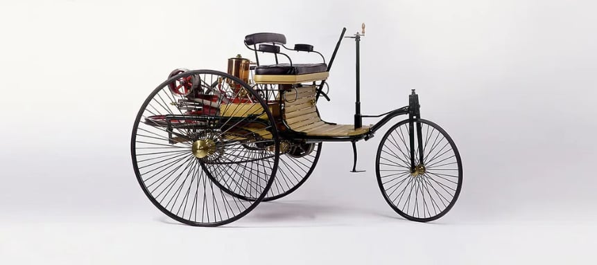 A vintage three-wheeled motor vehicle with thin metal spokes, wooden seats, and exposed mechanical parts, shown against a plain white background.