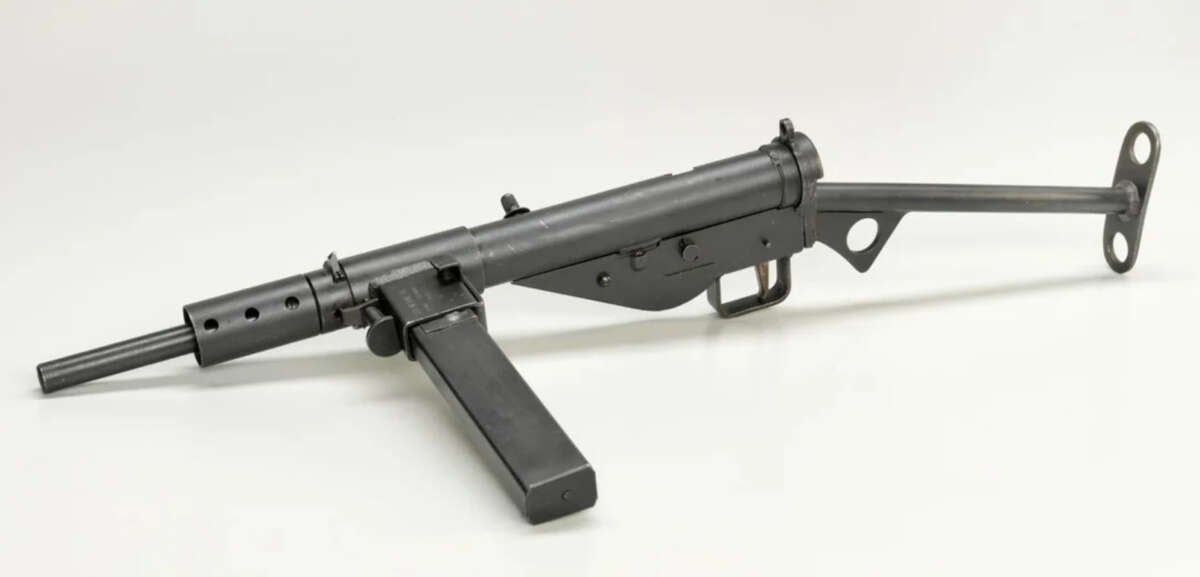 A Sten submachine gun with a metal frame, a side-mounted magazine, and a simple skeletal stock, displayed on a plain white background.
