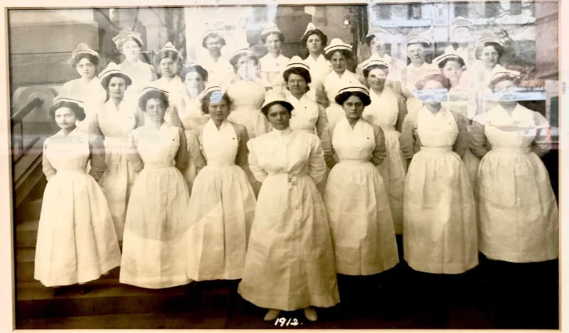 A black-and-white photo from 1912 shows a group of women in white nursing uniforms and hats, standing in rows on outdoor steps in front of a building.