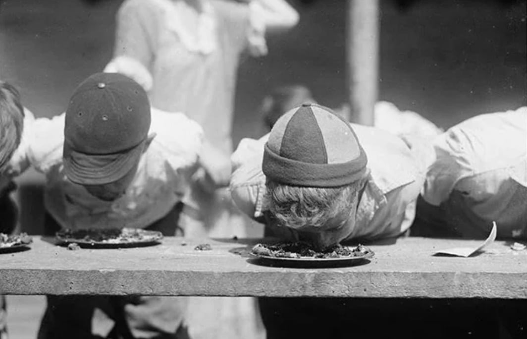 Two children with hats lean over a table, faces in their plates, appearing to compete in a pie-eating contest. A blurry figure stands in the background.