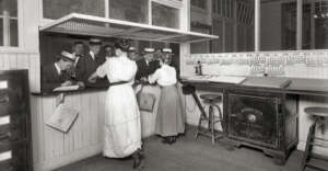 Black-and-white photo of men in hats standing at a counter, talking to two women in long skirts. The room has wood paneling, wire windows, a large safe, and an old-fashioned telephone on the counter.