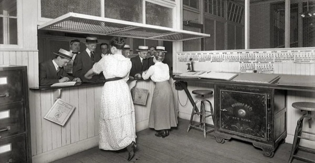 Black-and-white photo of men in hats standing at a counter, talking to two women in long skirts. The room has wood paneling, wire windows, a large safe, and an old-fashioned telephone on the counter.