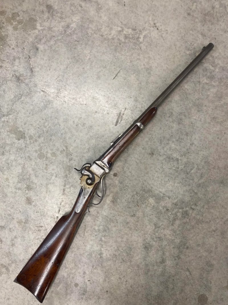 An antique lever-action rifle with a wooden stock and metal barrel is lying on a gray concrete surface. The rifle appears aged, showing some wear and patina on the wood and metal parts.